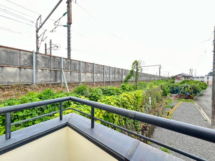 黒い手すりが設置されたバルコニーからの眺望です。周囲に高い建物はなく、開けた景色が広がっています。敷地に隣接して緑の植栽が配置され、奥には線路が通る環境となっています。