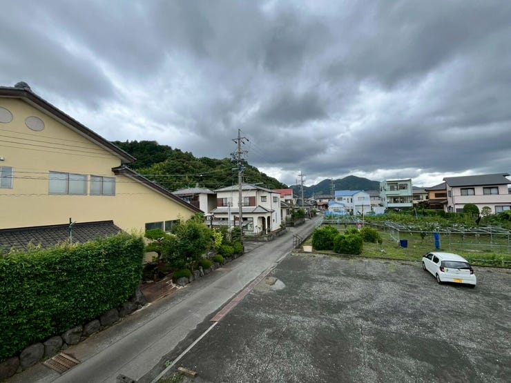 周囲に低層の住宅や空き地、緑豊かな山並みが広がる開放的な眺望です。手前には舗装された道路があり、視界を遮る高層の建物が周囲に少ないため、空が広く見渡せる環境となっています。