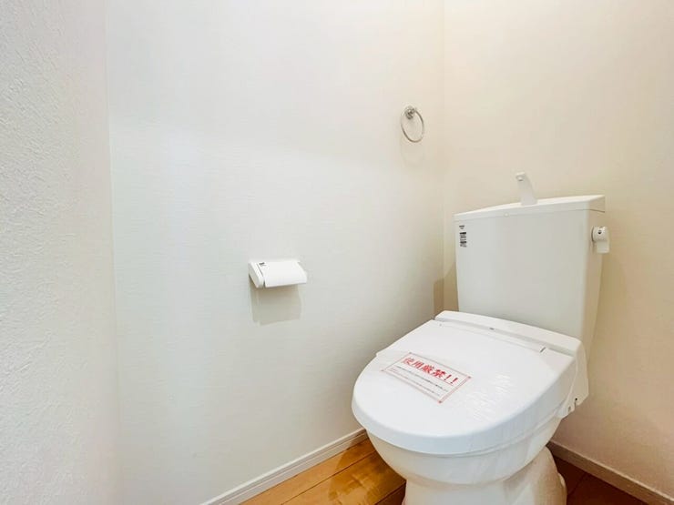 白を基調とした壁面に木目調の床材を合わせたトイレ空間です。手洗い機能付きのタンクと温水洗浄便座が一体となった衛生設備が設置されております。壁面にはタオル掛けやホルダーがあらかじめ配置された構造です。