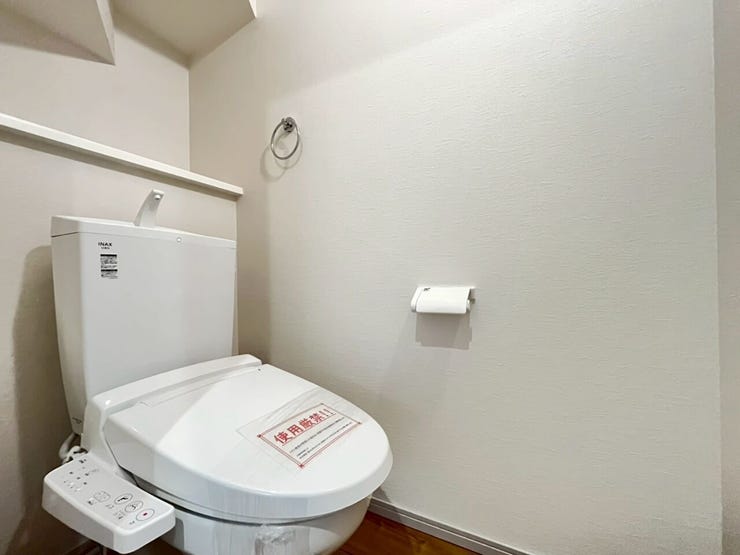 白を基調とした壁に木目調の床材を組み合わせたトイレ空間です。設備として手洗い器付きのタンクと、温水洗浄便座が設置されております。また便器の背面には小物を配置できる棚が設けられた構造となっております。