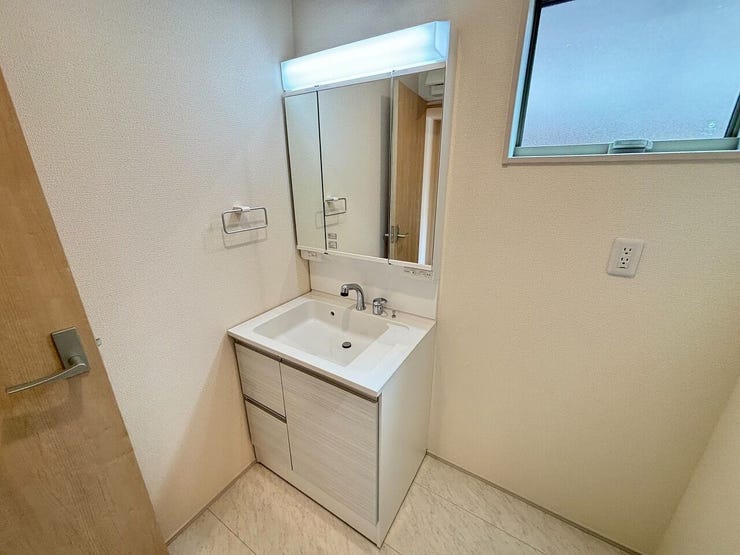 白を基調とした内装の洗面室です。洗面化粧台は三面鏡仕様となっており、下部には引き出しと開き扉の収納スペースが備わっています。壁面上部には窓が設けられており、自然光を取り入れつつ換気も行える設計です。