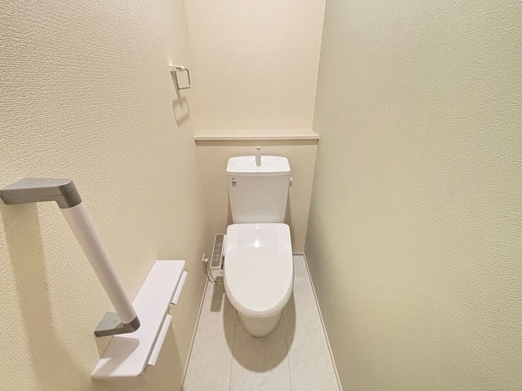 白を基調とした内装のトイレです。壁面に手すりが設置されており、立ち座りの動作に配慮された設計です。ペーパーホルダー上部や便器後方にはカウンターが設けられ、小物を配置するスペースが確保されています。