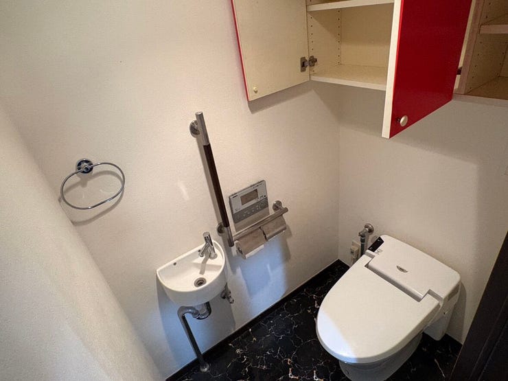 独立した手洗い場を備えた、ゆとりあるトイレ空間です。上部には赤い扉がアクセントの収納棚があり、ペーパー類もすっきり収まります。手すりも設置されているため、どなたでも安心してお使いいただける設計です。
