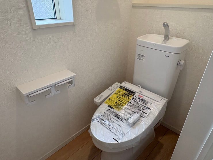 清潔感のある白を基調とした、温水洗浄機能付きの快適なトイレ。ペーパーホルダーが2つ設置されており、日々の使いやすさにも配慮されています。さりげないアクセントクロスが、品よく空間を彩るデザインです。