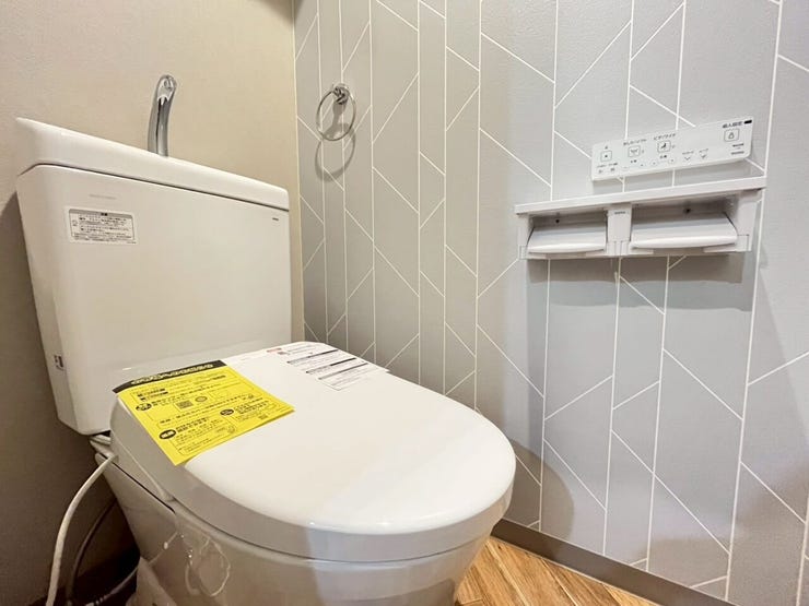 幾何学模様のアクセントクロスを採用したトイレ空間です。手洗い器付きの便器を設置し、壁面には温水洗浄便座の操作パネルと2連ペーパーホルダーを備えた、実用的な設備構成の内装となっております。