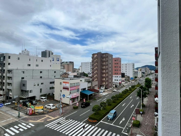 バルコニーからは市街地を見渡す景色が広がる住戸です。前面には植栽が整備された複数車線の道路が通り、周辺には商業ビルやマンションが立ち並んでいます。遠方の山並みを背景とした開放的な眺望です。