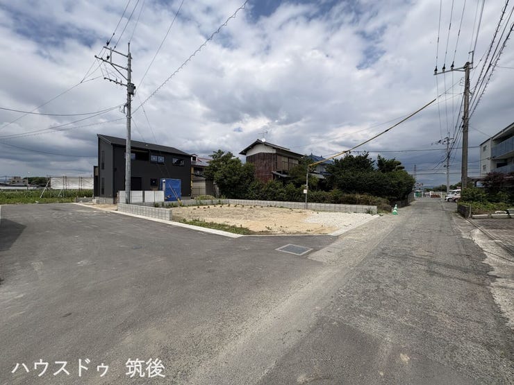 【前面道路含む現地写真】西側幅員6m公道と南側道路が交差する位置から撮影した、着工前の更地です。この角地風の敷地内に、図面通りの住戸や駐車スペースが完成します。