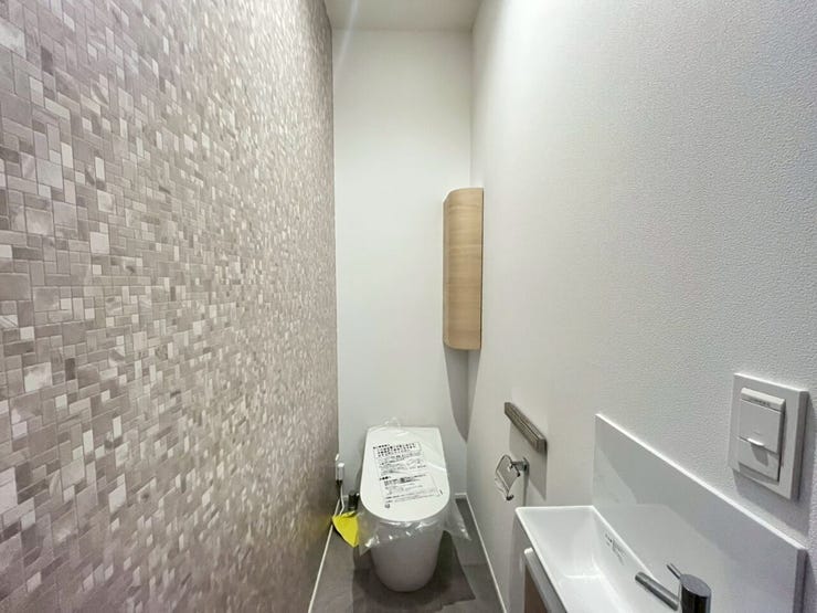 片側の壁にモザイクタイル調のクロスを採用したトイレです。独立した手洗い器と木目調のコーナー収納が設けられており、空間をすっきりと維持できる設計がなされています。