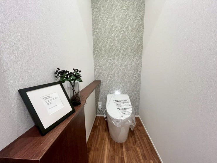 1階トイレ
木目調の床と植物柄のアクセントクロスを採用したトイレです。側面に木目調カウンターが設置されており、小物の配置スペースが確保された設計がなされています。