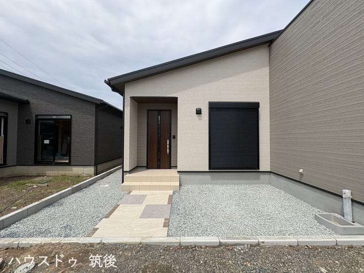 【片流れ屋根の平屋外観】シャープな印象の片流れ屋根を採用した平屋建ての外観です。木目調の玄関ドアへと続くアプローチが整えられており、手前の窓には防犯や防災にも役立つシャッターが標準装備されています。
