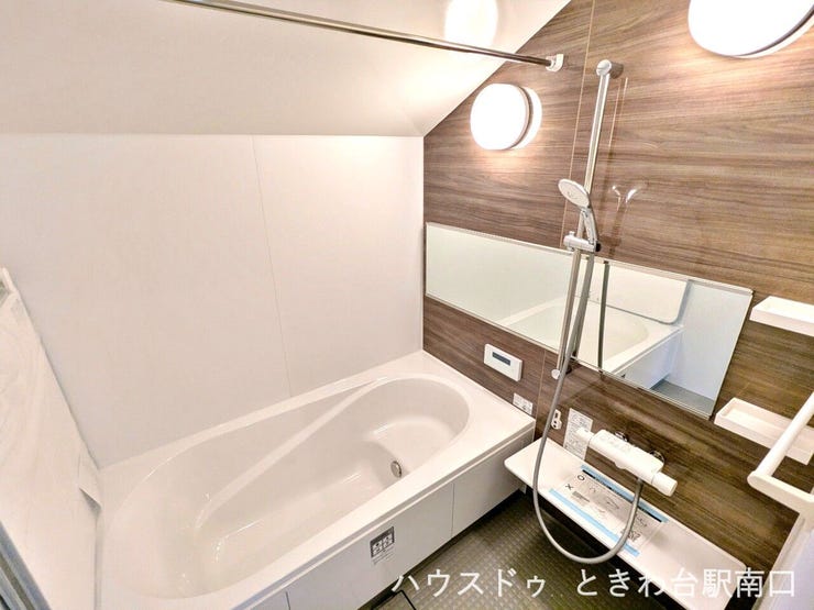 清潔感のあるホワイトの浴槽と、温かみのある壁面を組み合わせた浴室。機能的なシャワーヘッドや手すりも備わり、快適なバスタイムをサポートする配慮された設計となっています。
