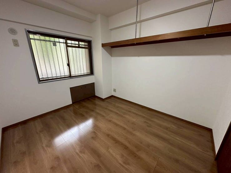 落ち着いたブラウンの床と白い壁のコントラストがすっきりとした印象の洋室。上部には長い吊り棚が設置されており、空間のアクセントと機能性を兼ね備えた構造デザインです。