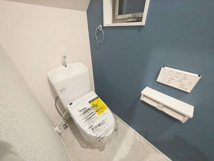 シックなブルーのアクセントクロスがおしゃれな印象を与えるトイレ空間。手洗い一体型で温水洗浄便座を装備しており、壁面リモコンやカウンター付きペーパーホルダー、小窓などの機能面も充実しています。