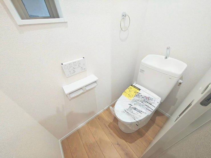 温かみのある木目調の床と清潔感のある白壁で仕上げたトイレ空間。手洗い一体型の温水洗浄便座に壁面リモコン、棚付きペーパーホルダーを完備し、上部の小窓で自然換気や採光も確保できます。