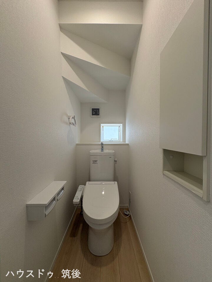 【小窓・収納棚付きトイレ】清潔感のある爽やかなトイレ空間です。自然光が入る小窓と換気扇を備え、通気性も良好。トイレットペーパーや小物を置ける便利な壁面収納棚も配置されています。