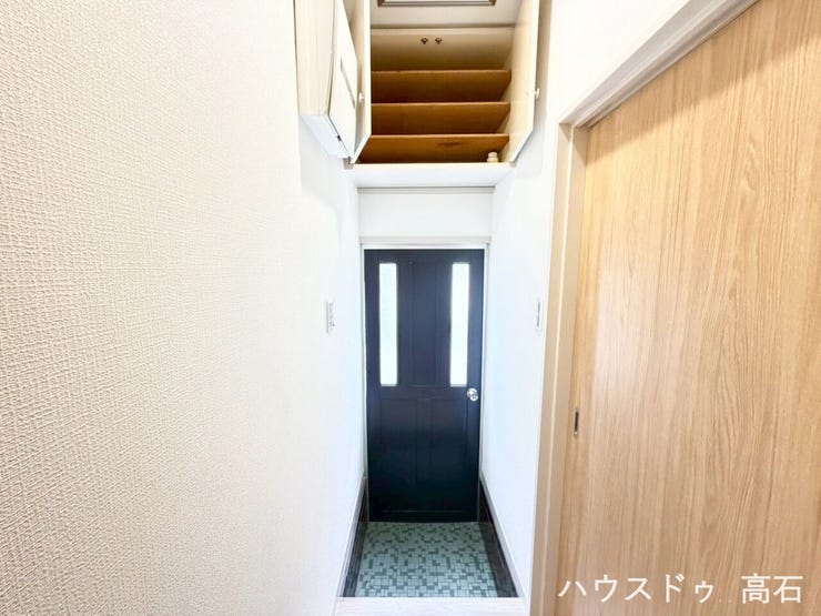 白を基調とした壁に黒のドアが映えるスタイリッシュな玄関。上部にはデッドスペースを有効活用した収納棚を備え、空間を無駄なく使える設計です。